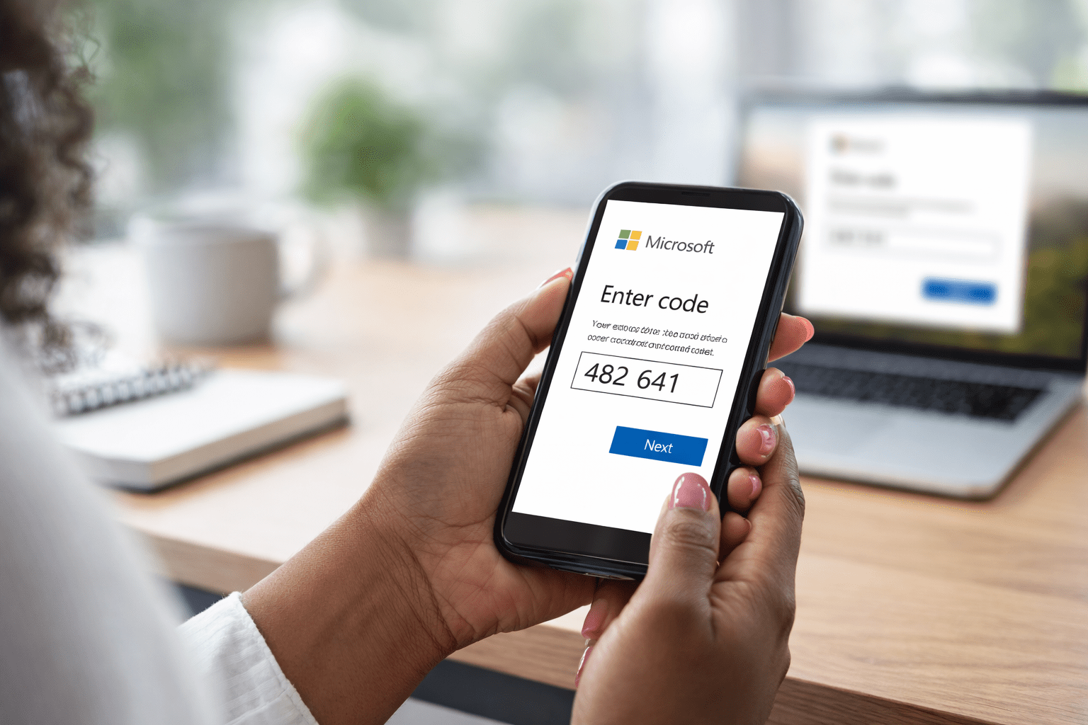 A woman with painted nails holds a smartphone displaying a Microsoft verification code in a bright office, with a laptop blurred in the background.