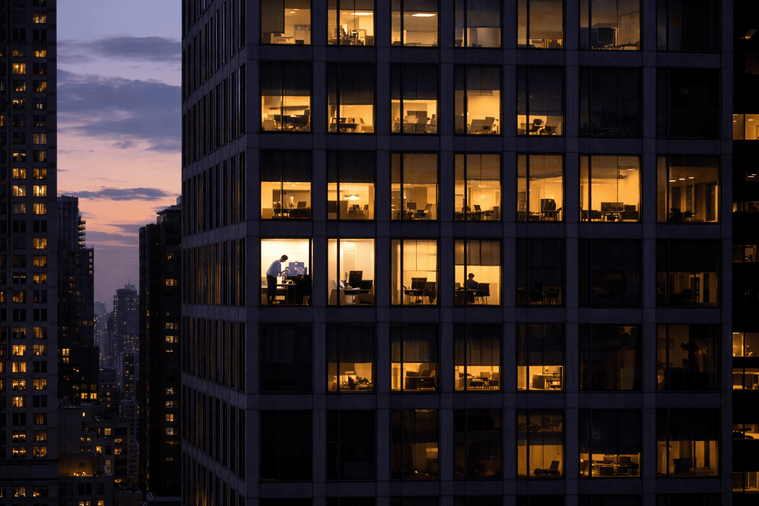 Tall New York City office building at dusk with some windows lit and others dark, showing different scenes inside: employees calmly working at desks, coworkers troubleshooting a computer, and empty offices after hours.