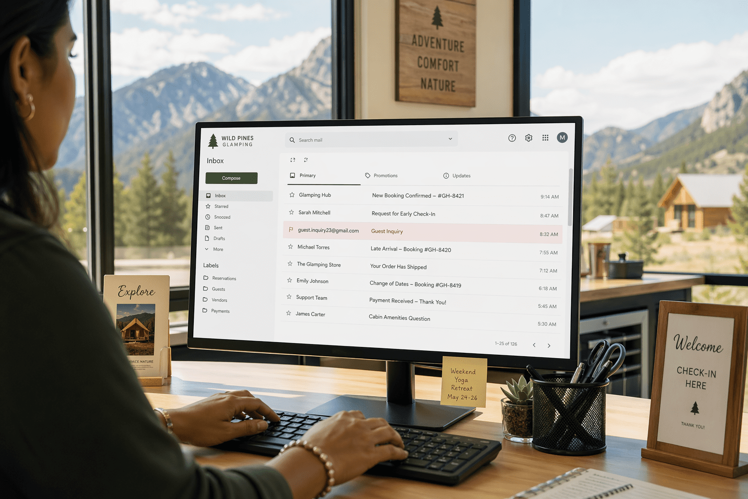 A woman’s hands hover over a keyboard at a bright, modern glamping reservations desk, as a computer screen displays an email inbox with a suspicious guest inquiry highlighted, suggesting a potential phishing attempt.